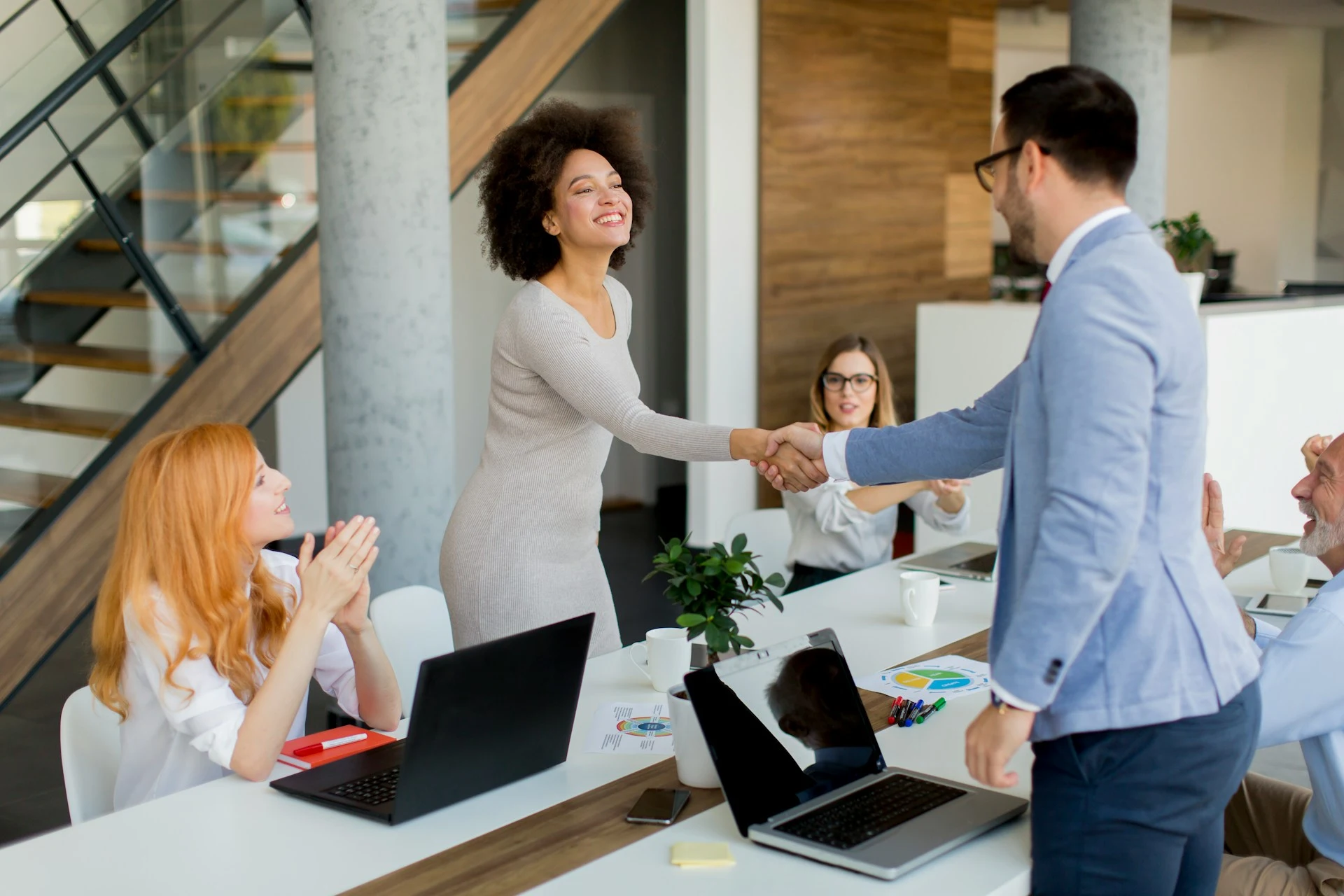 A room full of business people smiling and clapping. A man and the woman in the centre are shaking hands.