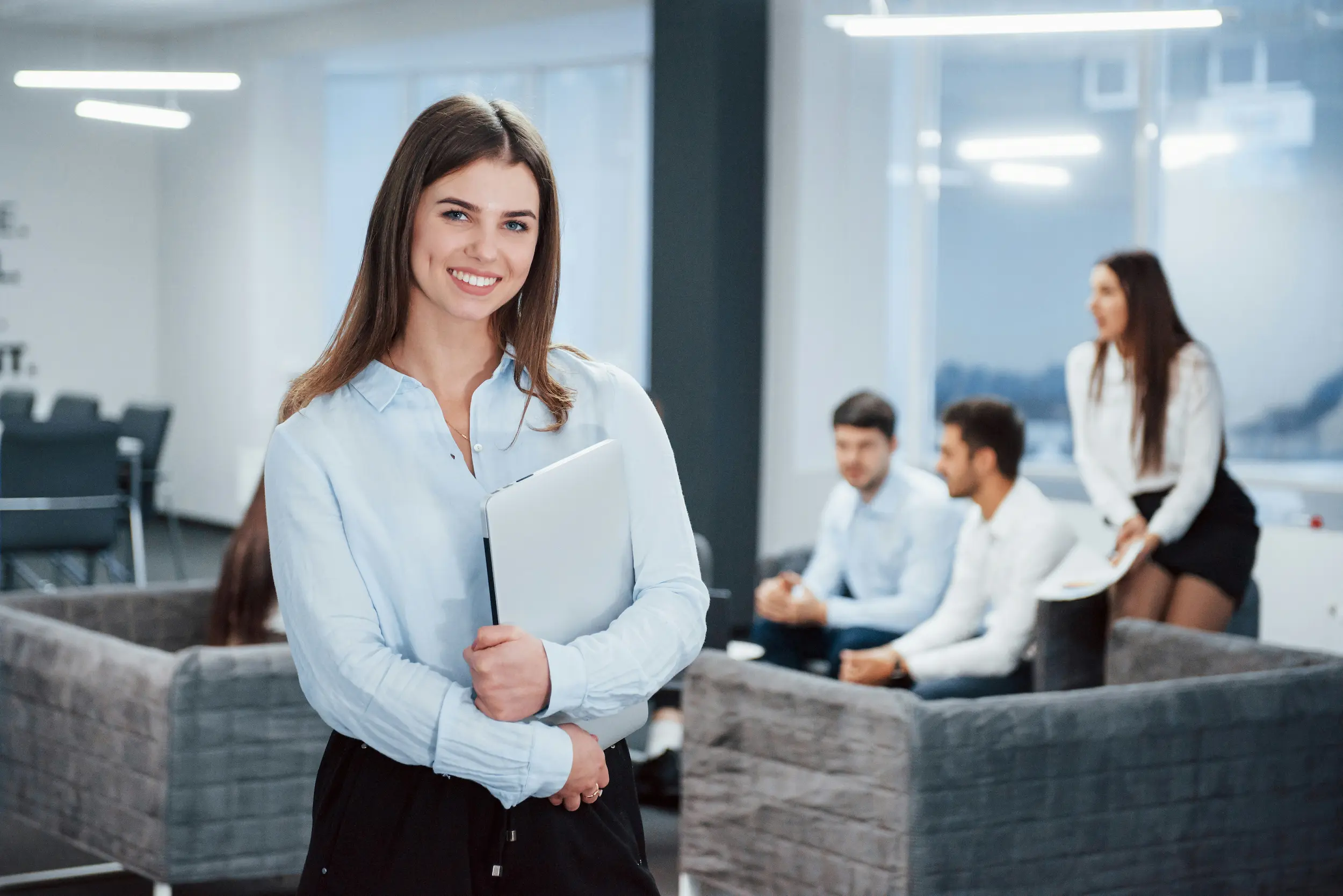  A temp worker standing in the office holding her laptop while a meeting is ongoing behind her.